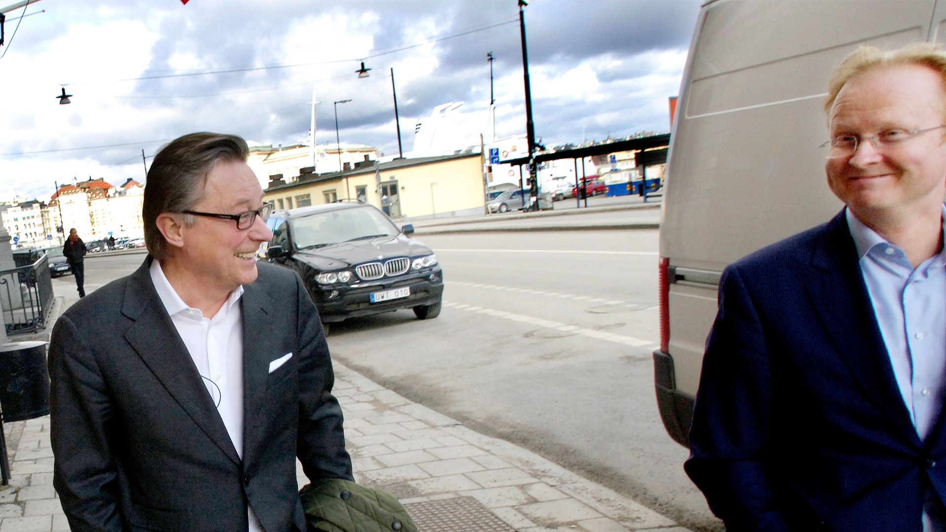Två män i affärskläder står på en parkering. Mannen till vänster bär glasögon och mörkblå kavaj. Mannen till höger bär ljusblå skjorta under mörkblå kavaj. I bakgrunden syns bilar och byggnader under en molnig himmel.