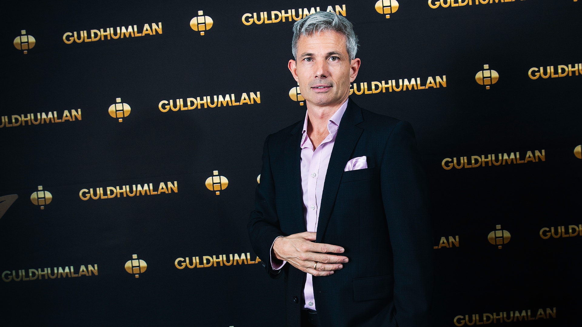 Man i svart kostym och rosa skjorta framför en mörk bakgrund med Guldhumlan-loggor.