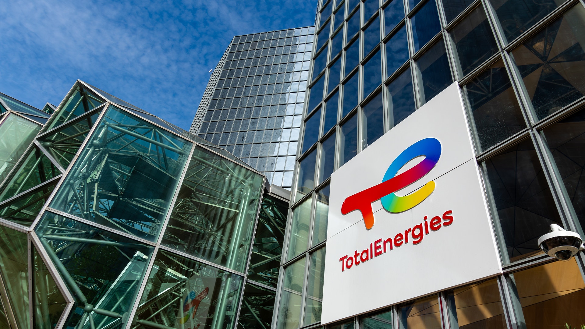 Skylt med TotalEnergies logotyp på en glasfasad, sedd underifrån mot blå himmel.