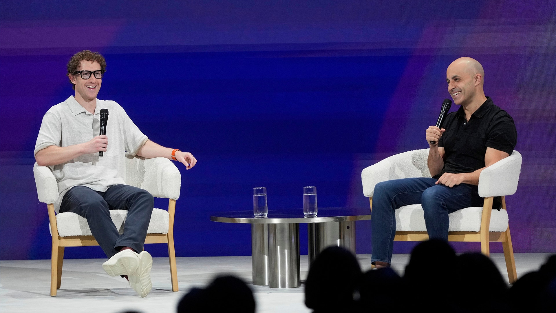 Två män sitter i stolar på en scen och samtalar framför en publik mot en blå bakgrund.