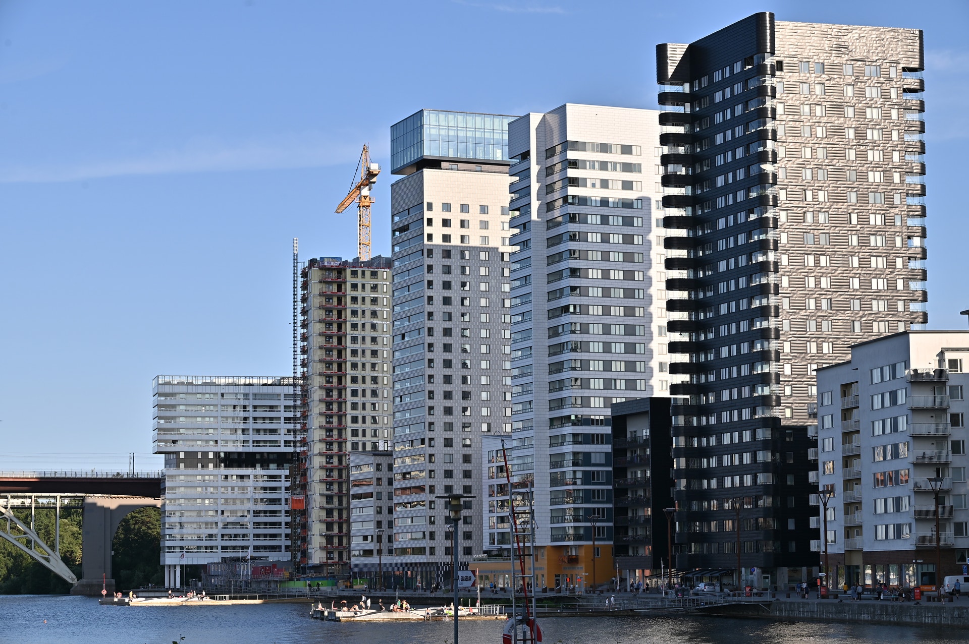 Moderna höghus längs en vattenkant under blå himmel. Flera bostadsbyggnader i olika höjder och färger, från vita till mörka fasader, med en byggkran synlig mellan husen.