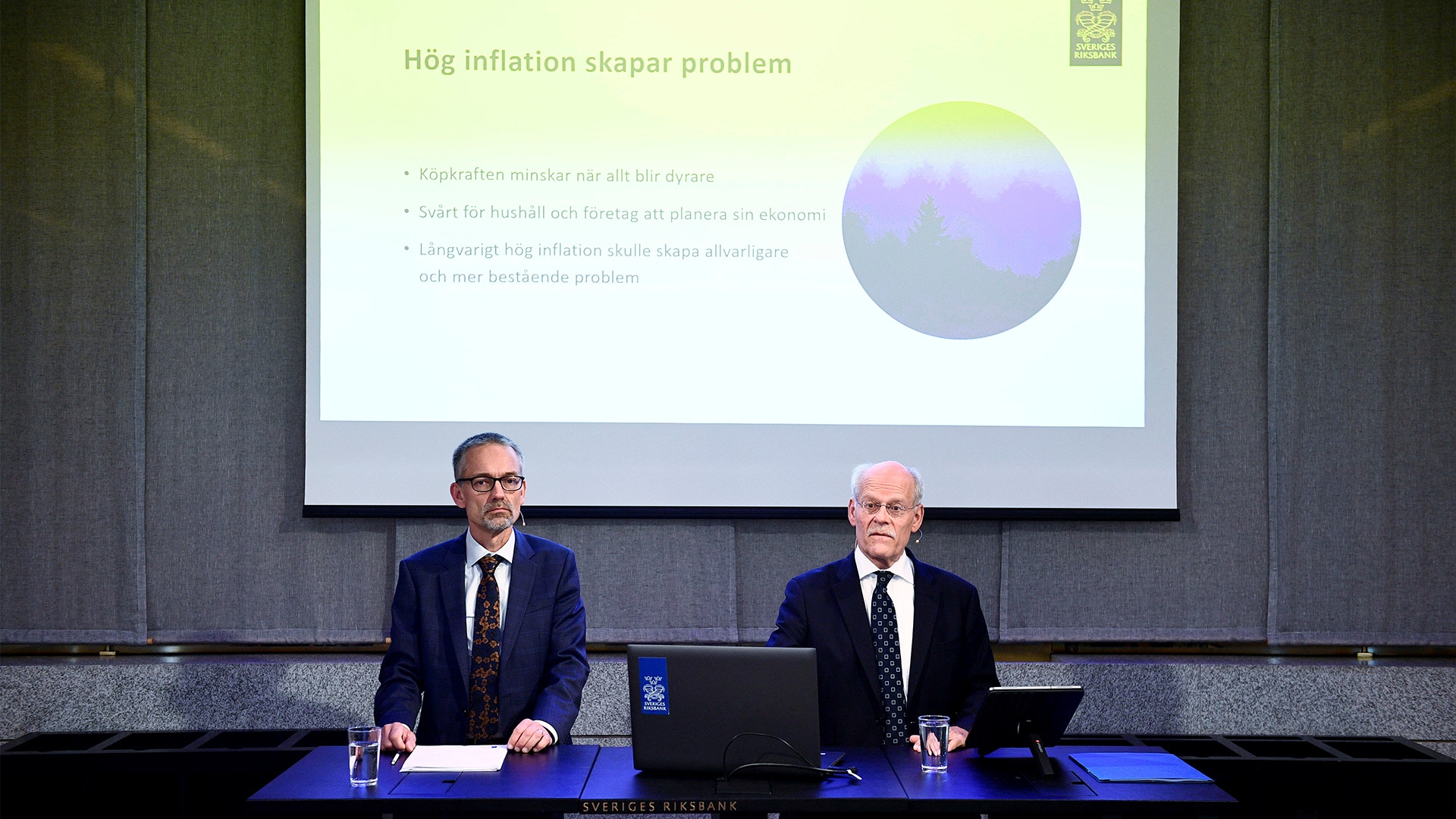 Två män i kostym sitter vid ett bord under en presentation om inflation från Riksbanken.