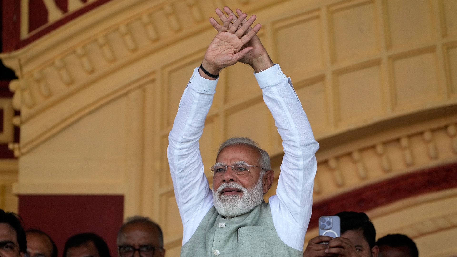 En man i vit skjorta höjer sina sammanfogade händer över huvudet i en segergäst framför en prydlig arkitektonisk bakgrund.