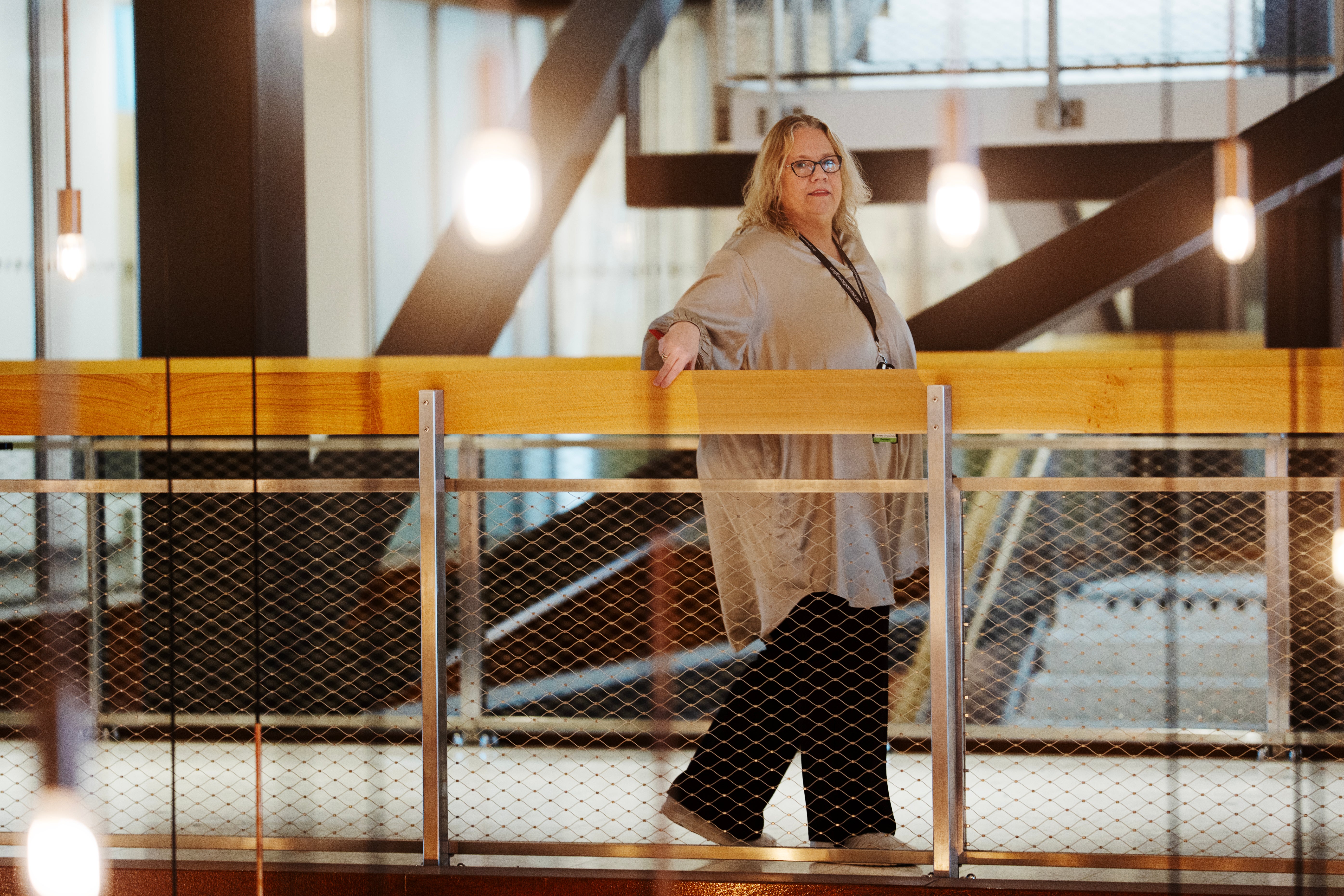 En kvinna med glasögon står vid ett räcke i ett modernt inomhusutrymme med träbalkar och hängande lampor.