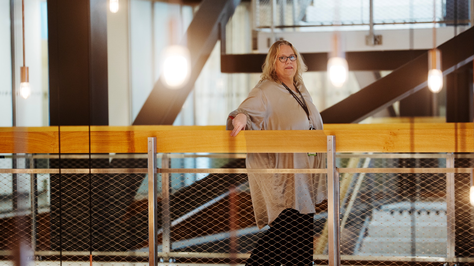 En kvinna med glasögon står vid ett räcke i ett modernt inomhusutrymme med träbalkar och hängande lampor.