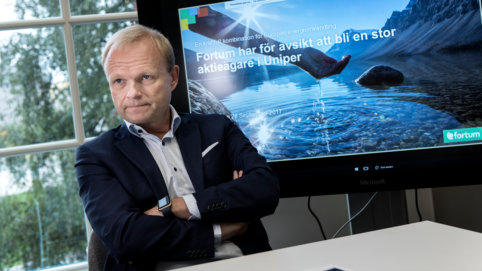 Man i mörkblå kostym sitter vid skrivbord med armarna korsade. I bakgrunden syns en stor skärm med en presentation.