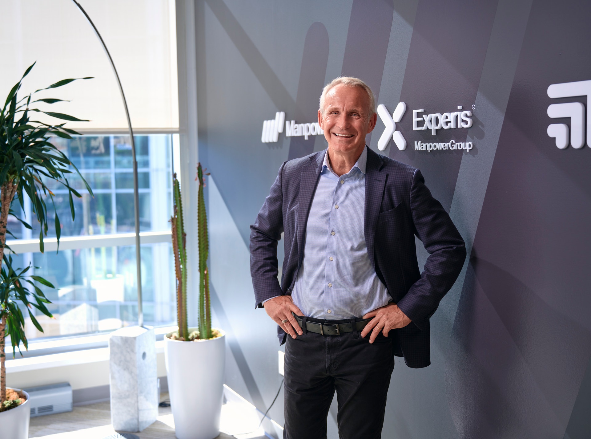 Leende man i kavaj står med händerna i sidorna framför en vägg med ManpowerGroups och Experis logotyper.