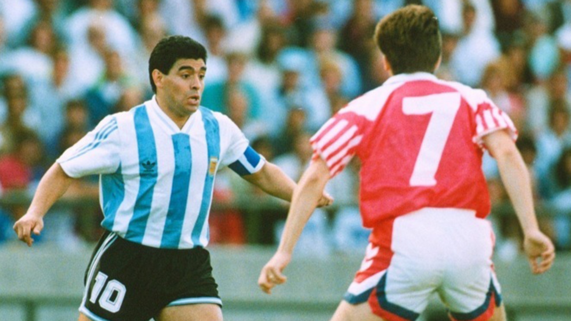 Fotbollsspelare i Argentinas blå-vita randiga tröja med nummer 10 spelar mot motspelare i röd tröja under en match med publik i bakgrunden.