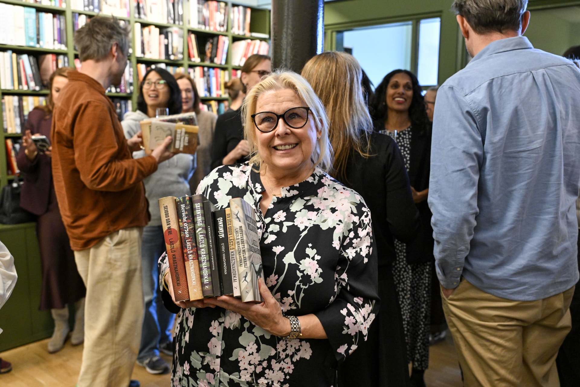 En leende kvinna med vit hår och glasögon håller flera böcker i ett bibliotek. I bakgrunden ses andra människor som bläddrar bland böcker på hyllor.