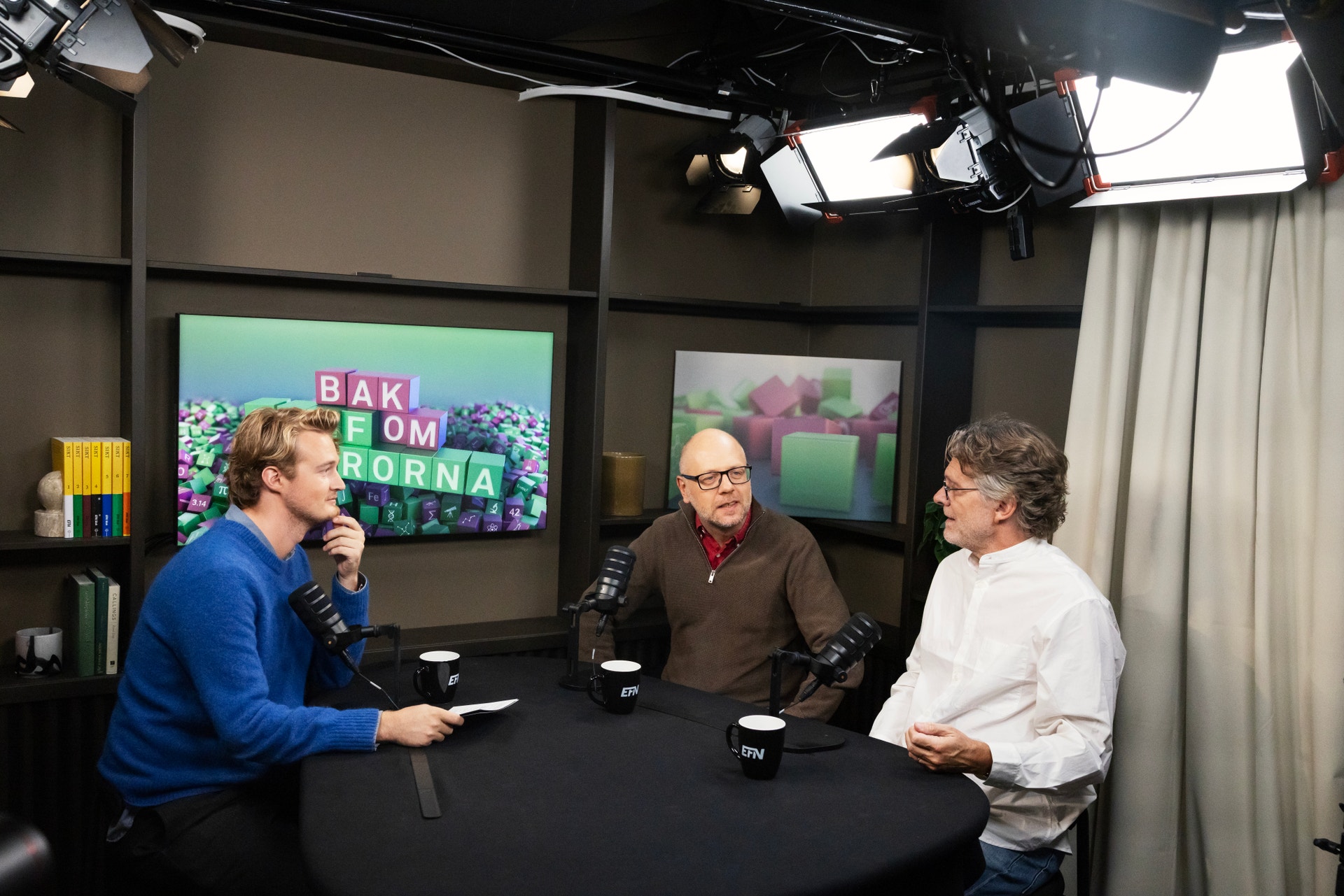 Tre män sitter vid ett runt bord med mikrofoner och samtalar i en podcaststudio.