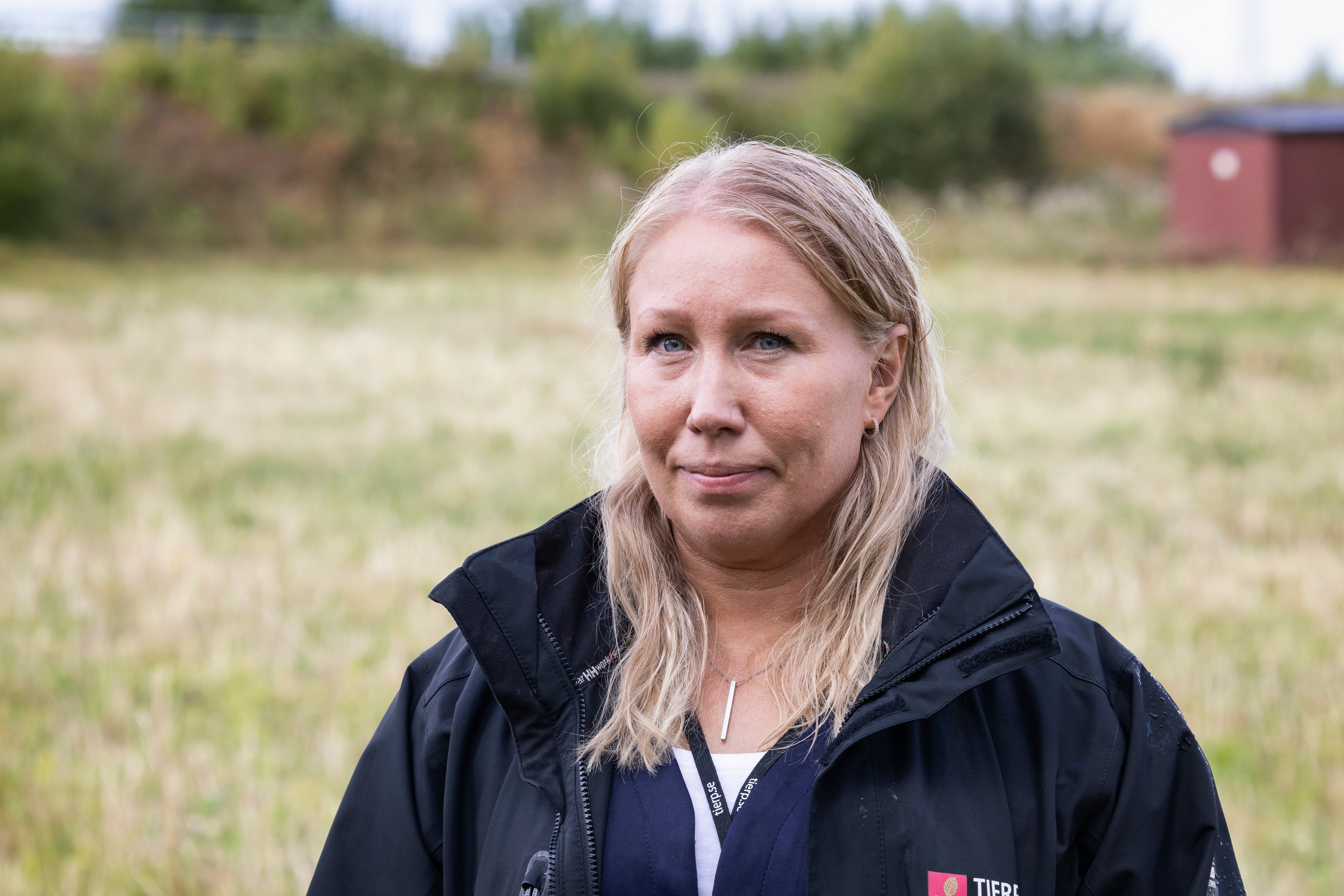 Porträtt av en kvinna med långt blont hår som står på en jordbruksmark. Hon bär en svart jacka och tittar mot kameran. I bakgrunden syns gröna fält, träd och en röd byggnad.