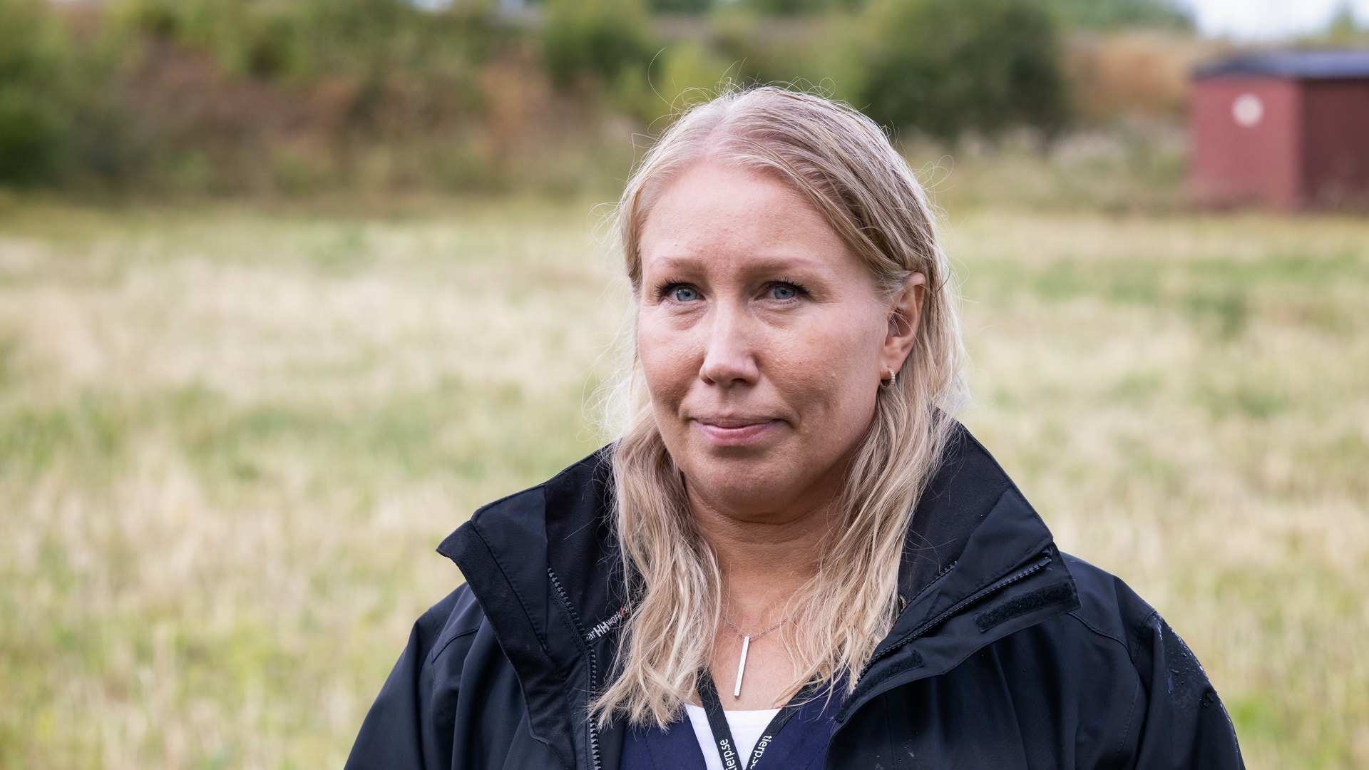 Porträtt av en kvinna med långt blont hår som står på en jordbruksmark. Hon bär en svart jacka och tittar mot kameran. I bakgrunden syns gröna fält, träd och en röd byggnad.