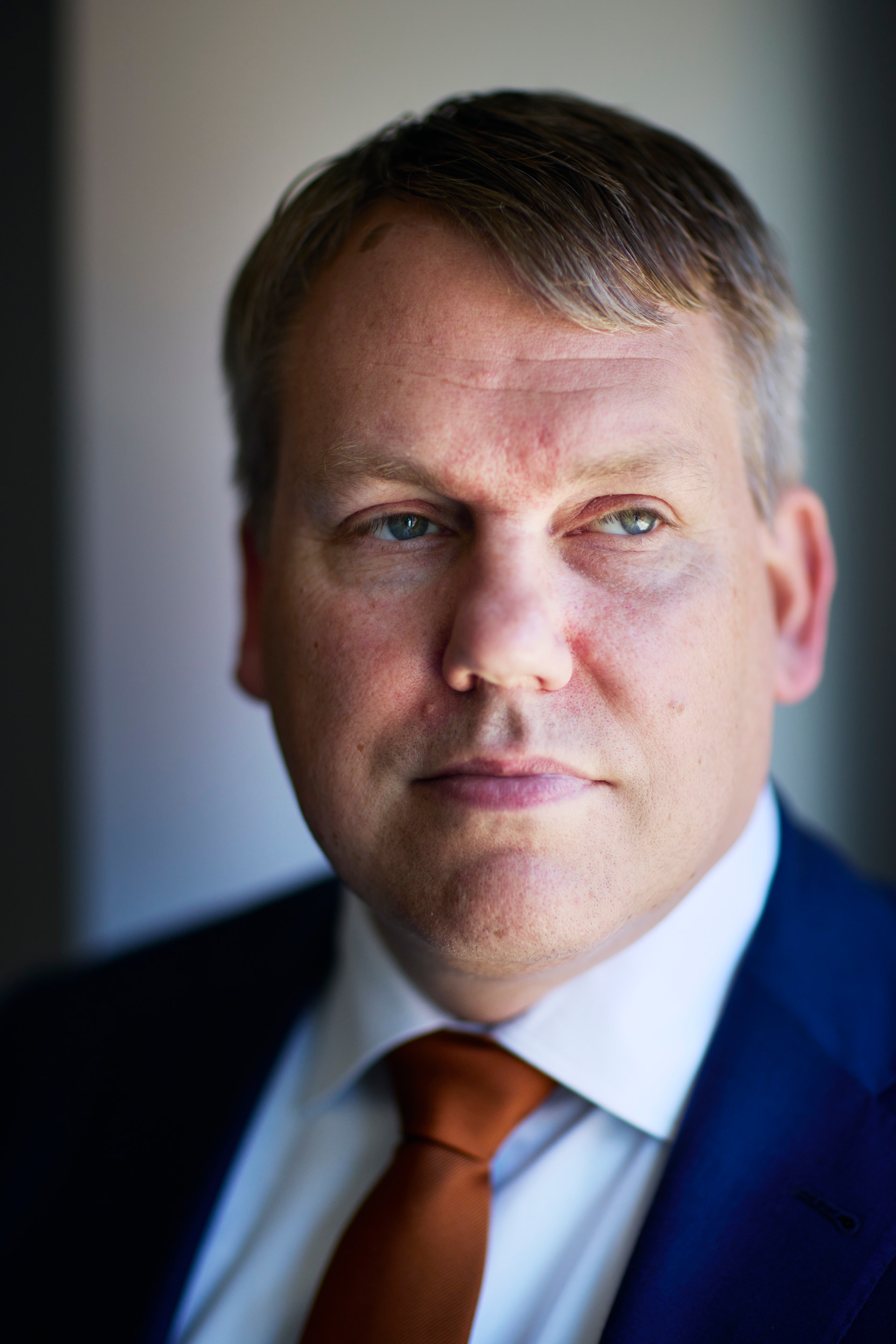 Porträtt av en man i mörk kostym, vit skjorta och orange slips mot neutral bakgrund.