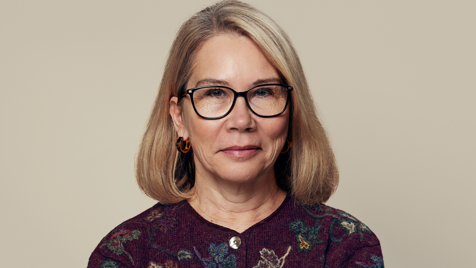 Porträtt av en kvinna med glasögon och långt brunt hår mot en beige bakgrund.