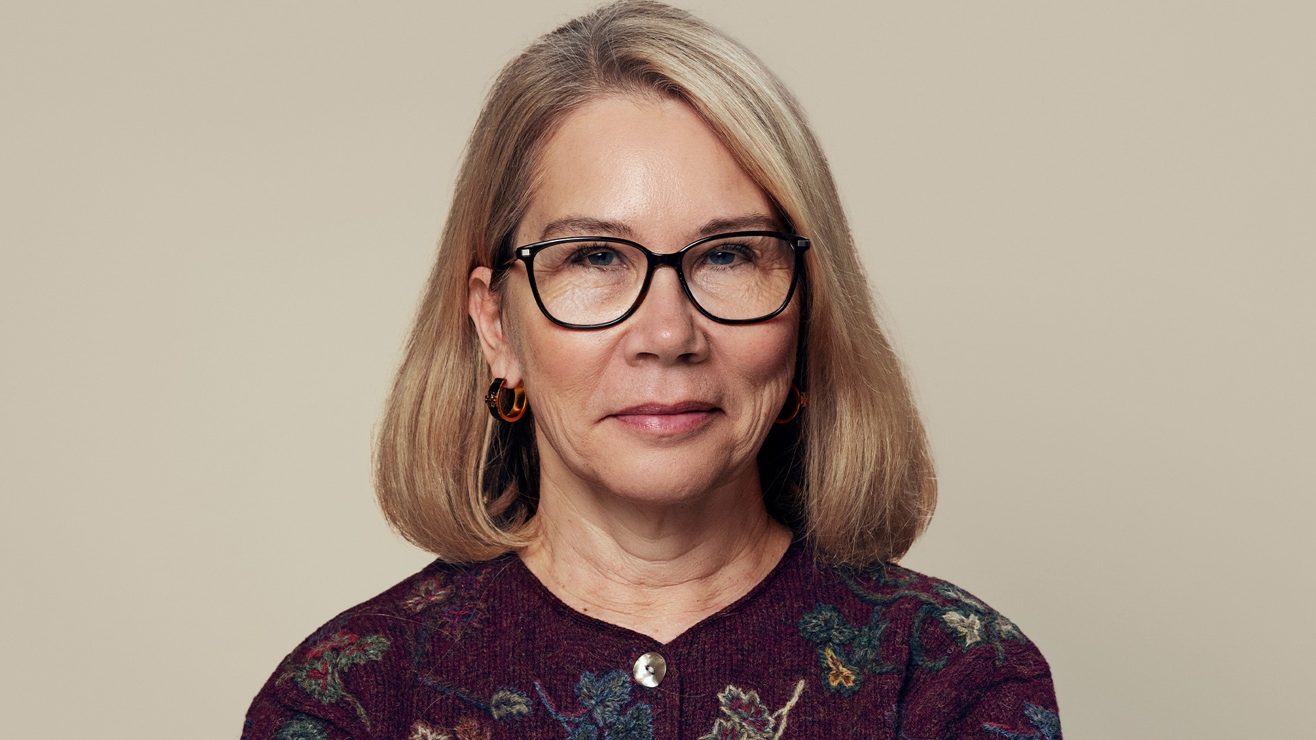 Porträtt av en kvinna med glasögon och långt brunt hår mot en beige bakgrund.