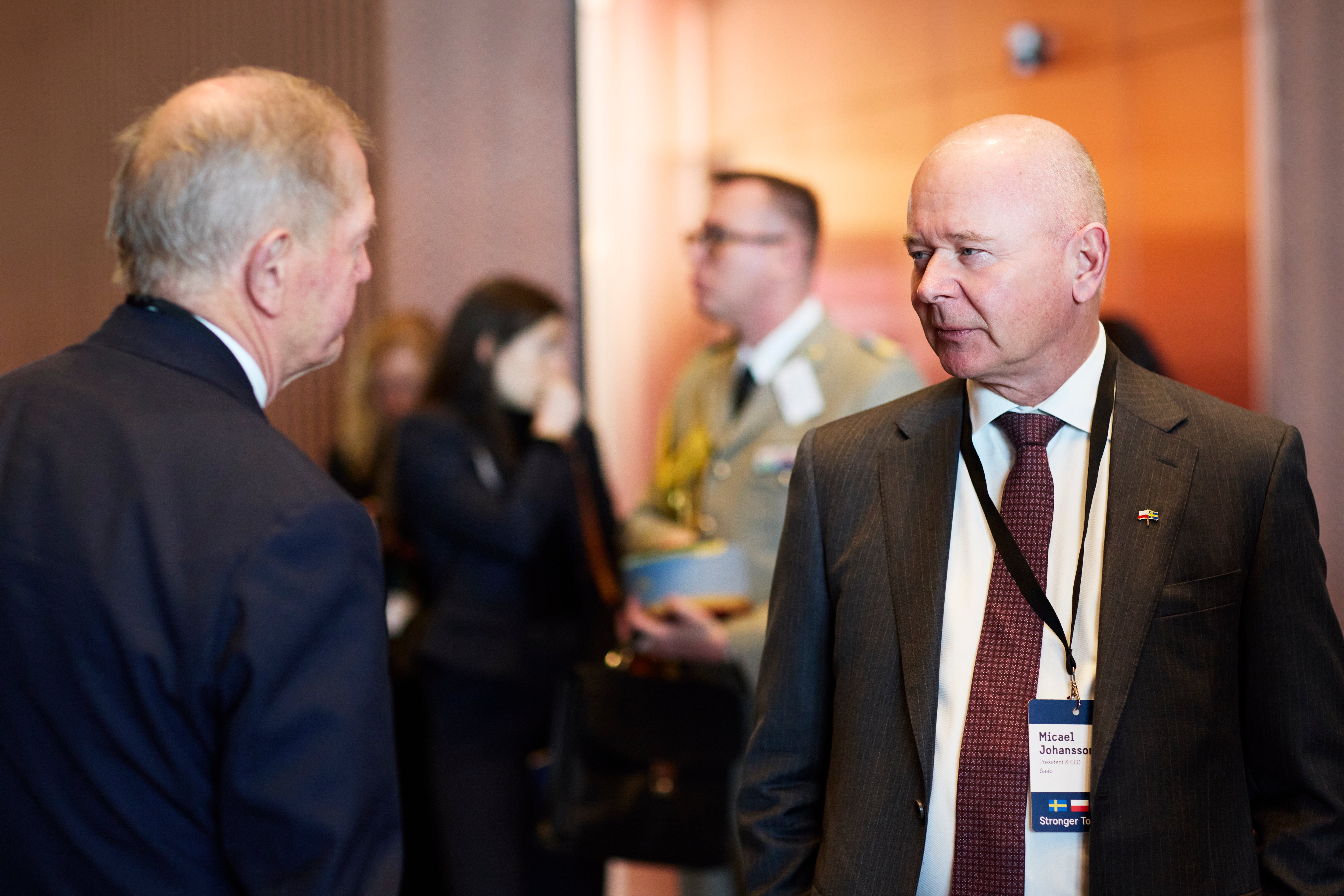 Två män i kostym samtalar vid en formell event inomhus.