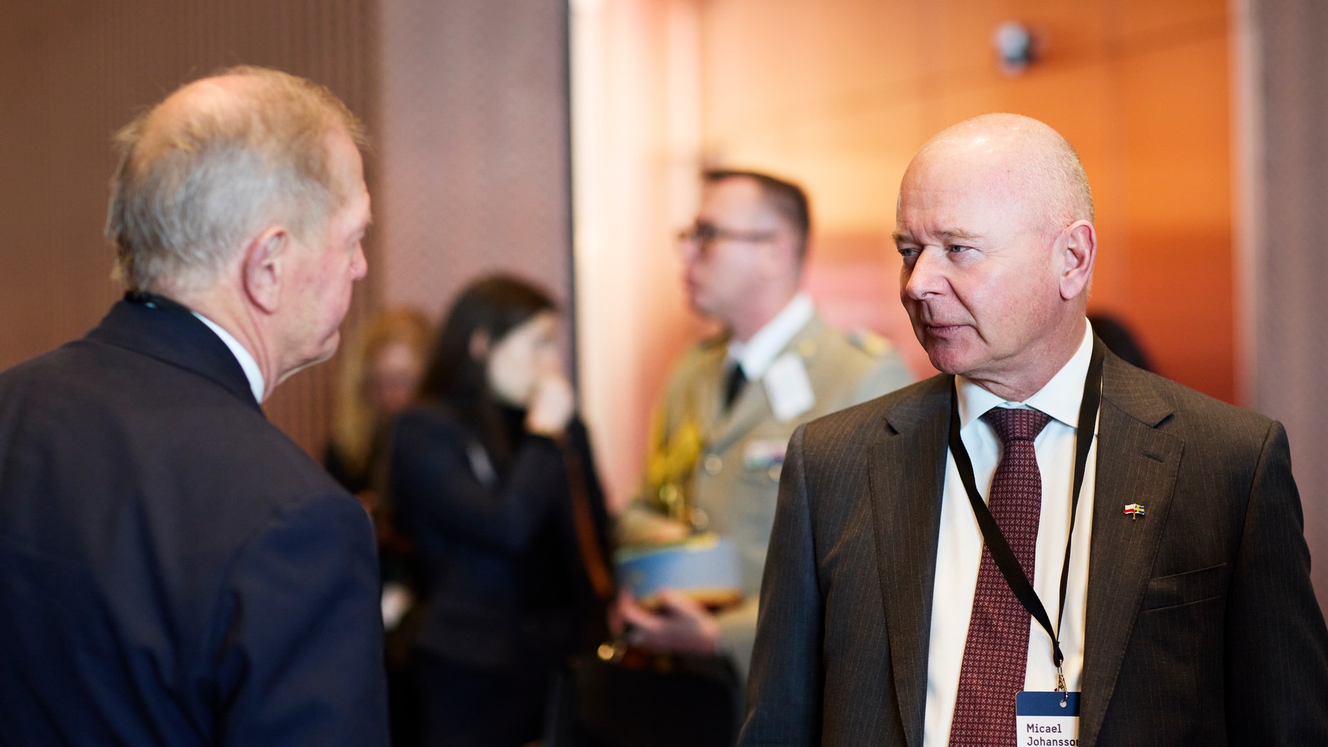 Två män i kostym samtalar vid en formell event inomhus.
