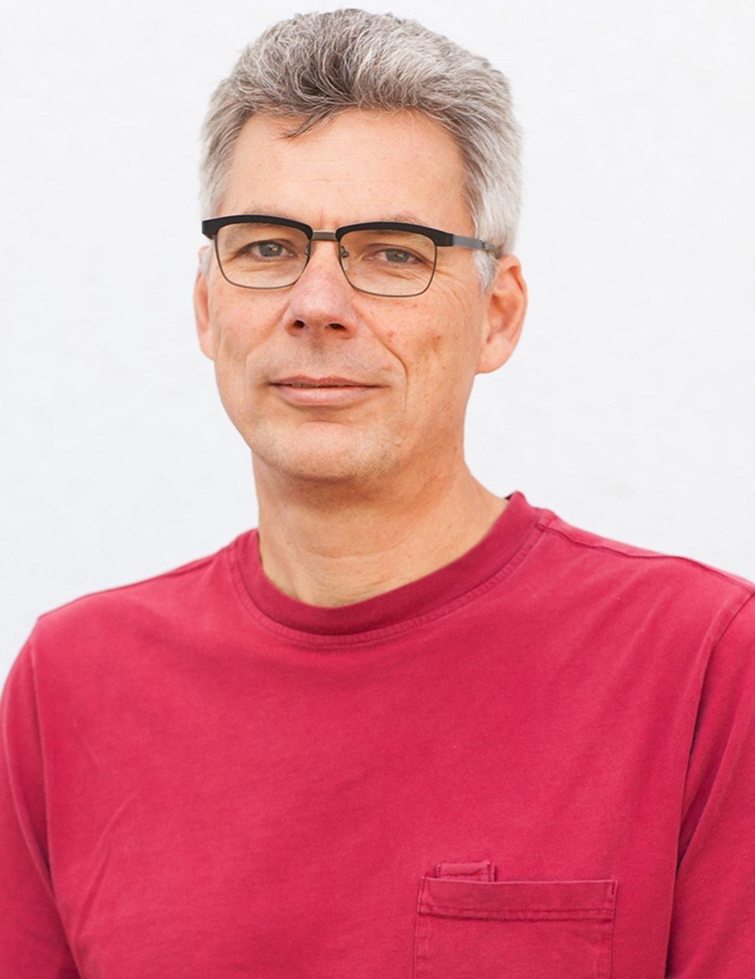 Porträtt av en man med grått hår och glasögon, iklädd en röd t-shirt mot vit bakgrund.