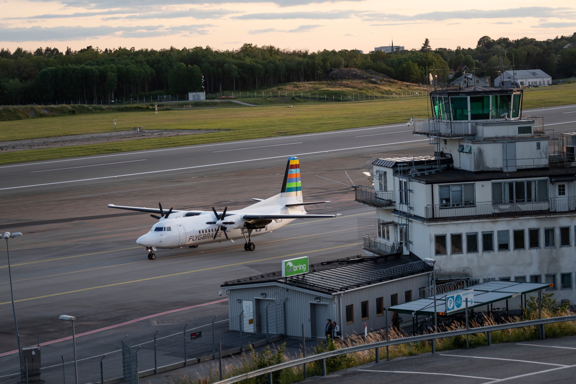 Ett turbopropdrivet passagerarflygplan står på landningsbanan vid en flygplats. I förgrunden syns en kontrolltorn och flygplatsbyggnader. I bakgrunden ses gröna ängar och barrskog.