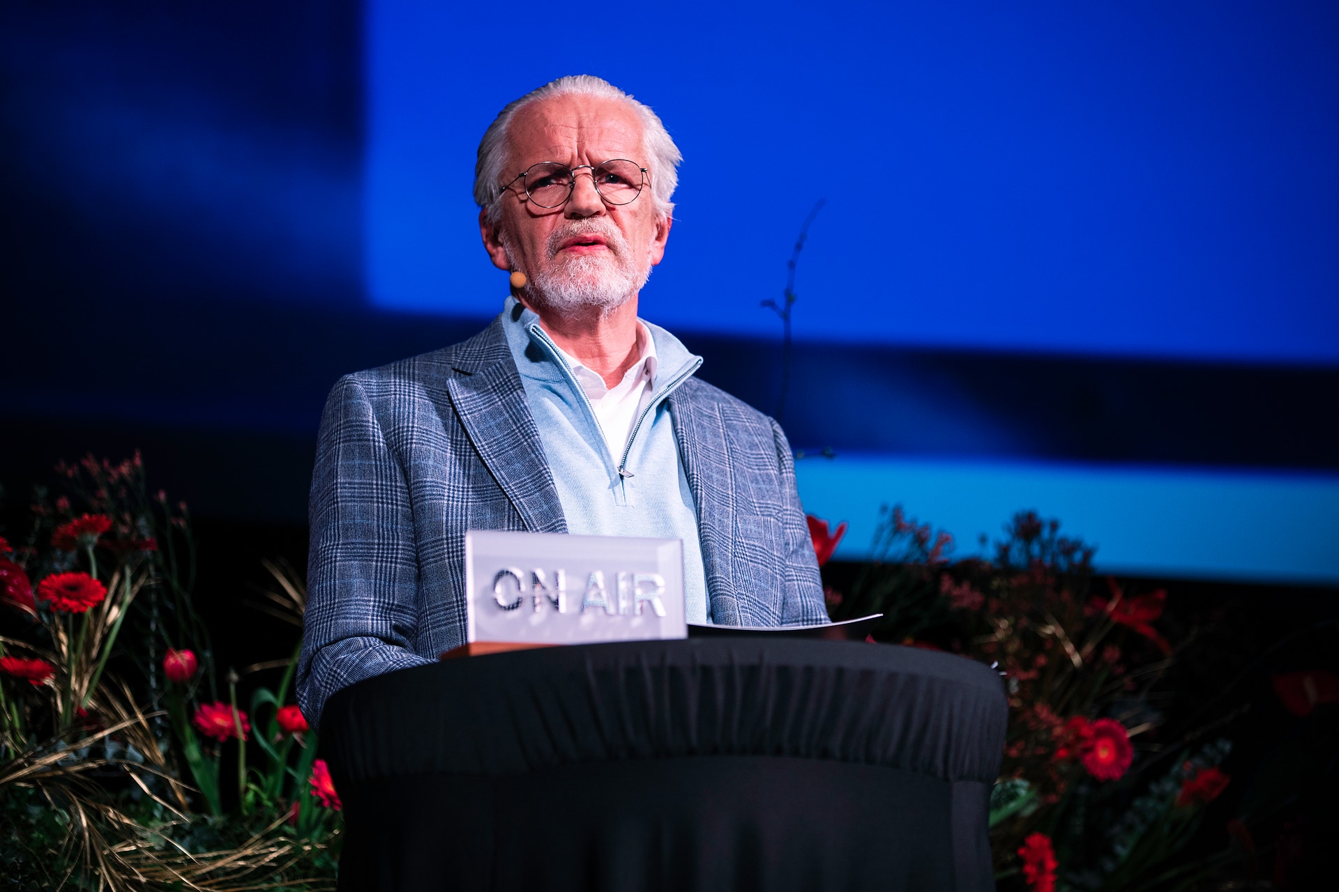 En äldre man i glasögon och grå kavaj står vid ett podium med ON AIR-skylt framför en blå bakgrund med röda blommor.