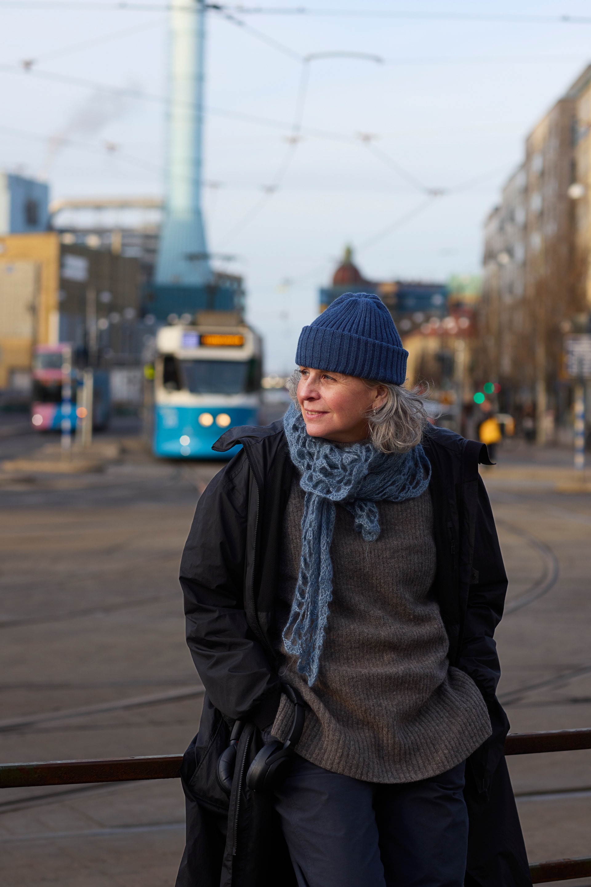 Person i mörkblå mössa och vinterkläder står på en stadsgata med en blå spårvagn i bakgrunden.