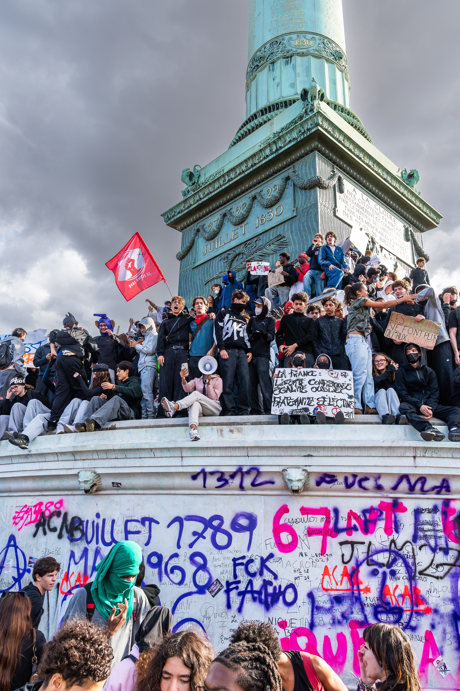 Många unga människor samlade vid en hög staty på en cirkelrund sockel i en stad, med plakat och en röd flagga. Sockeln är täckt av klotter.