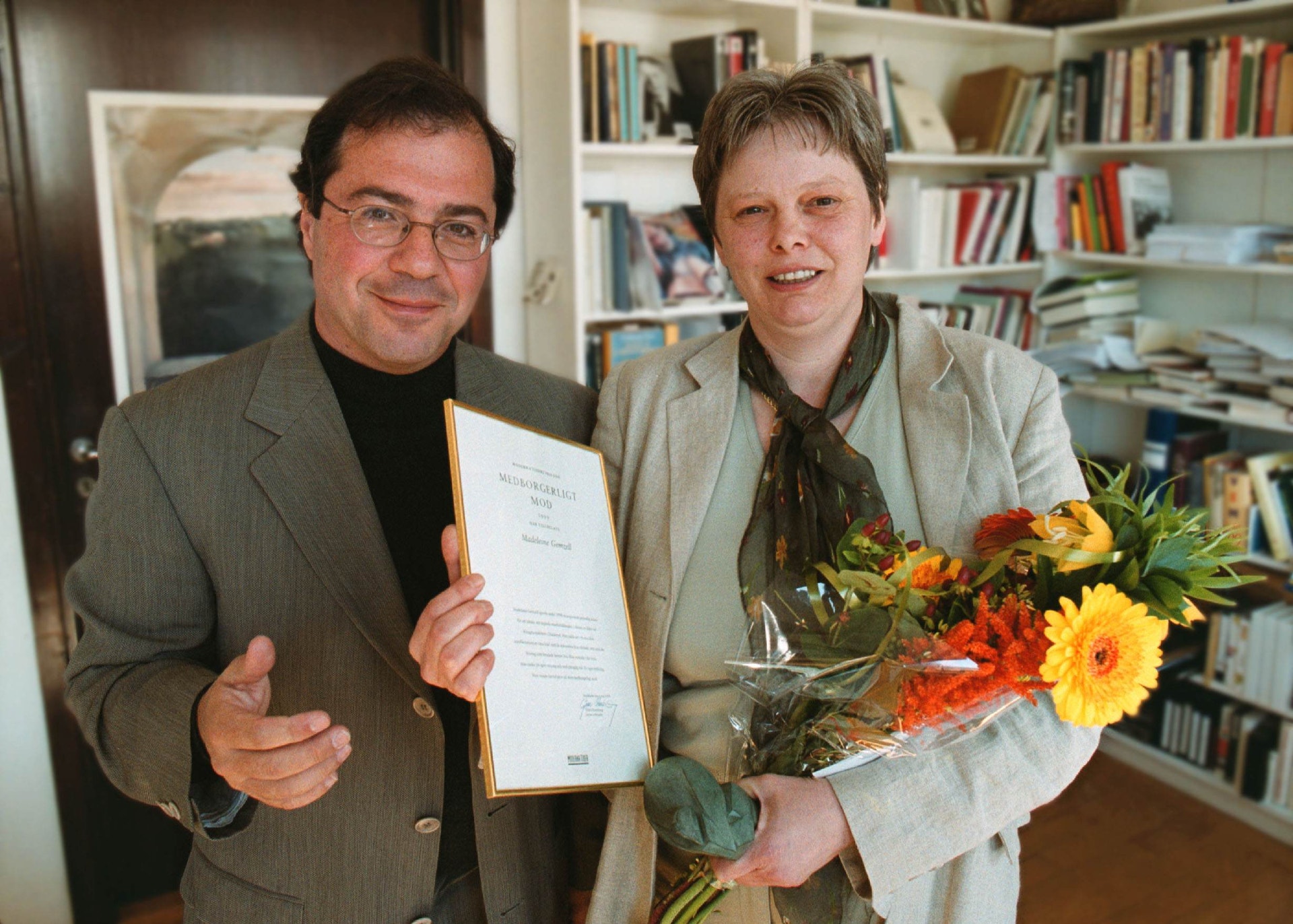 Två personer står tillsammans inomhus framför en bokhylla. Personen till vänster håller upp ett diplom eller certifikat, och personen till höger håller en färgglad blomsterbukett.