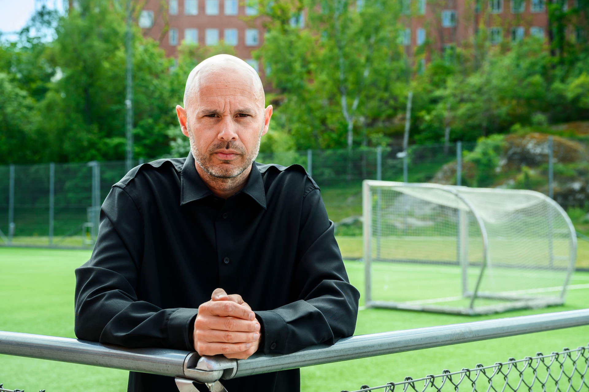 Man i svart jacka står vid ett fotbollsplansstaket. I bakgrunden syns ett fotbollsmål, grönt gräs och träd.