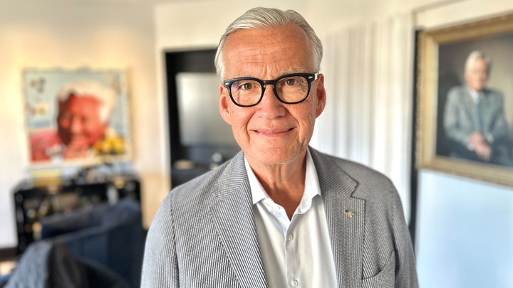 Porträtt av en äldre man med glasögon och grå hår, klädd i ljus skjorta och grå kavaj, fotograferad inomhus.