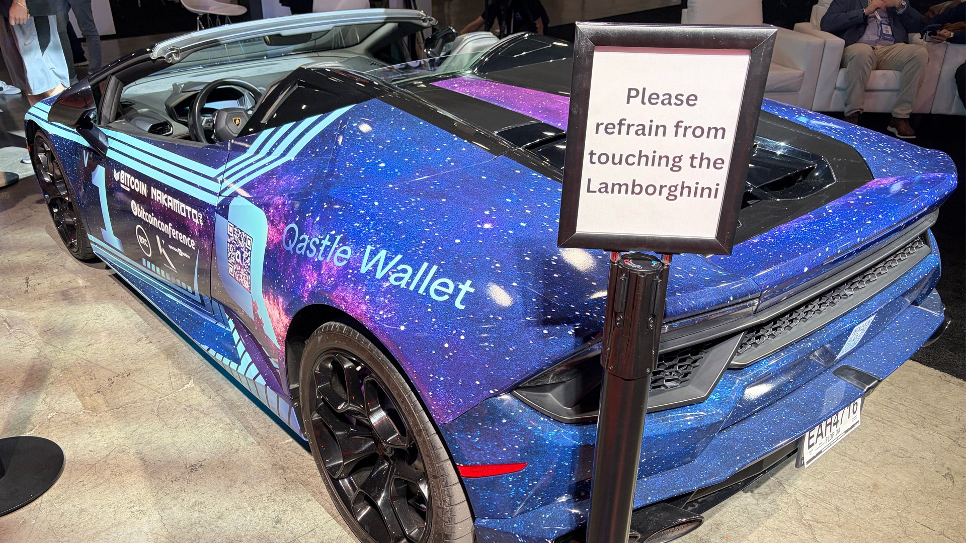 En blå Lamborghini med galaxmönster i lila och blå färger på en utställning. En skylt framför bilen instruerar besökare att inte röra vid bilen.