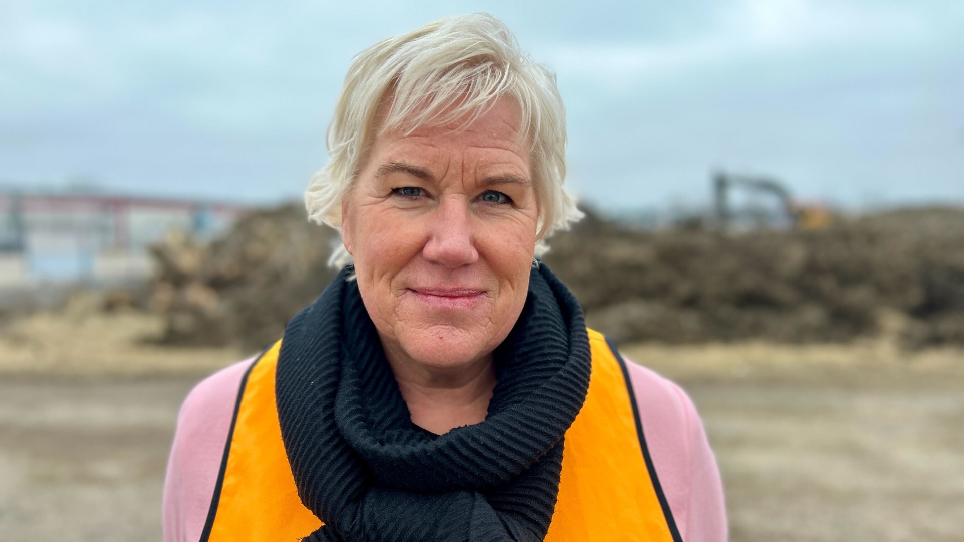 Porträtt av en kvinna med kort blont hår som bär en rosa tröja och en orange väst. Hon står utomhus på en byggarbetsplats och ler mot kameran.