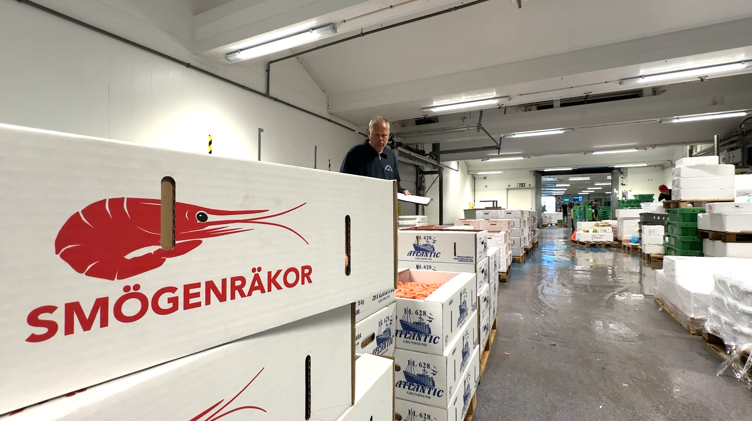 Produktionsanläggning för Smögenråkor med företagets röda logotyp på vägg, arbetare och packade produkter synliga i bakgrunden.