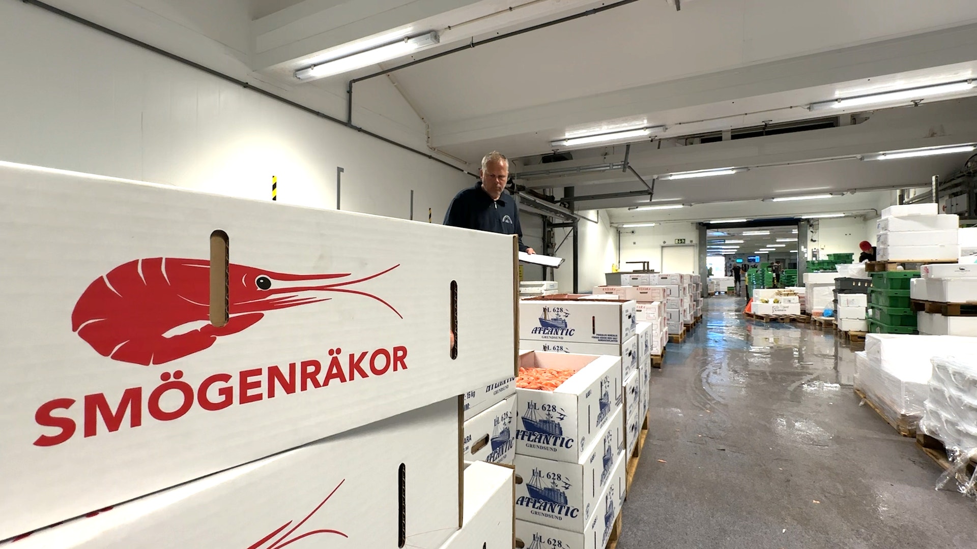 Produktionsanläggning för Smögenråkor med företagets röda logotyp på vägg, arbetare och packade produkter synliga i bakgrunden.