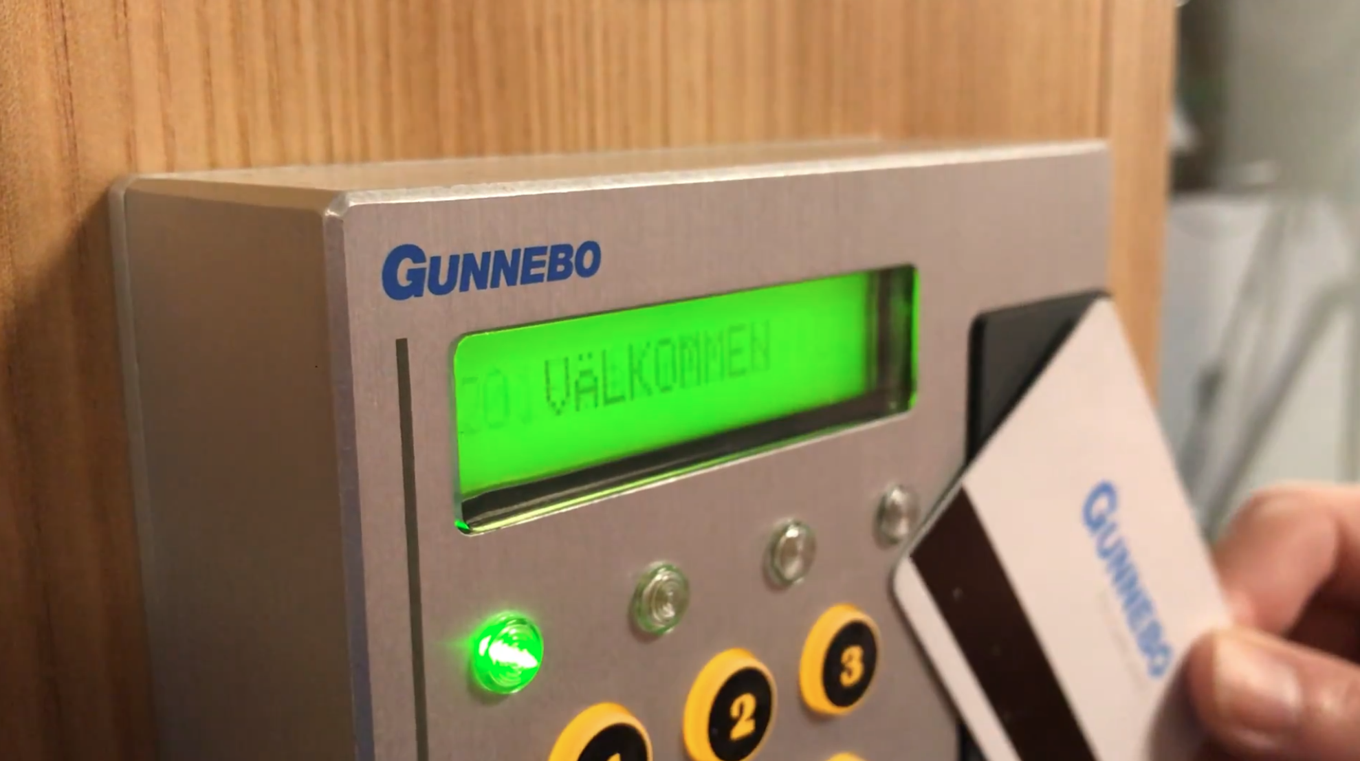 Närbild av en Gunnebo-säkerhetsenhet med grön LCD-display som visar texten Välkommen, tillsammans med gröna och gula indikatorer samt kontrollknappar.