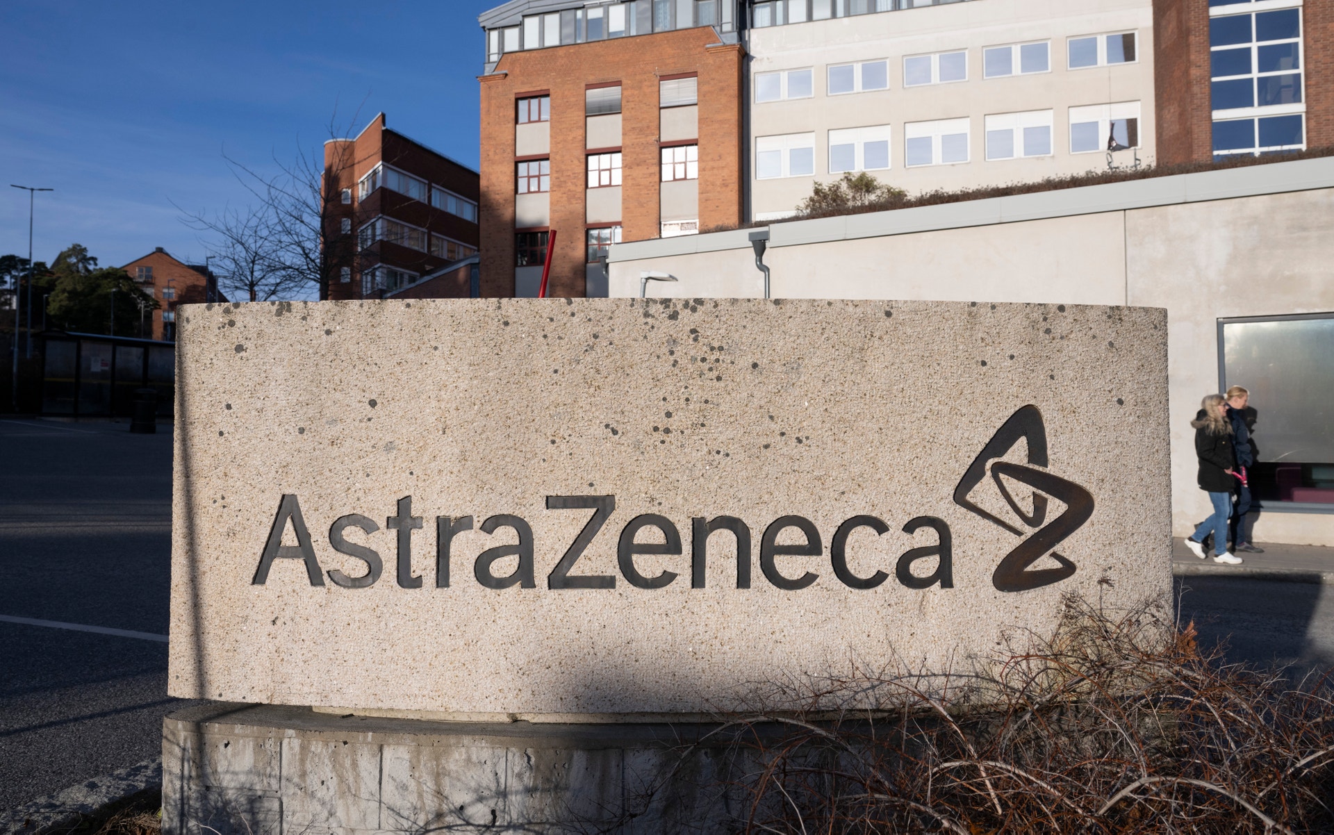 Betongvägg med AstraZeneca-logotypen vid företagets anläggning, med kontorsbyggnader i bakgrunden.