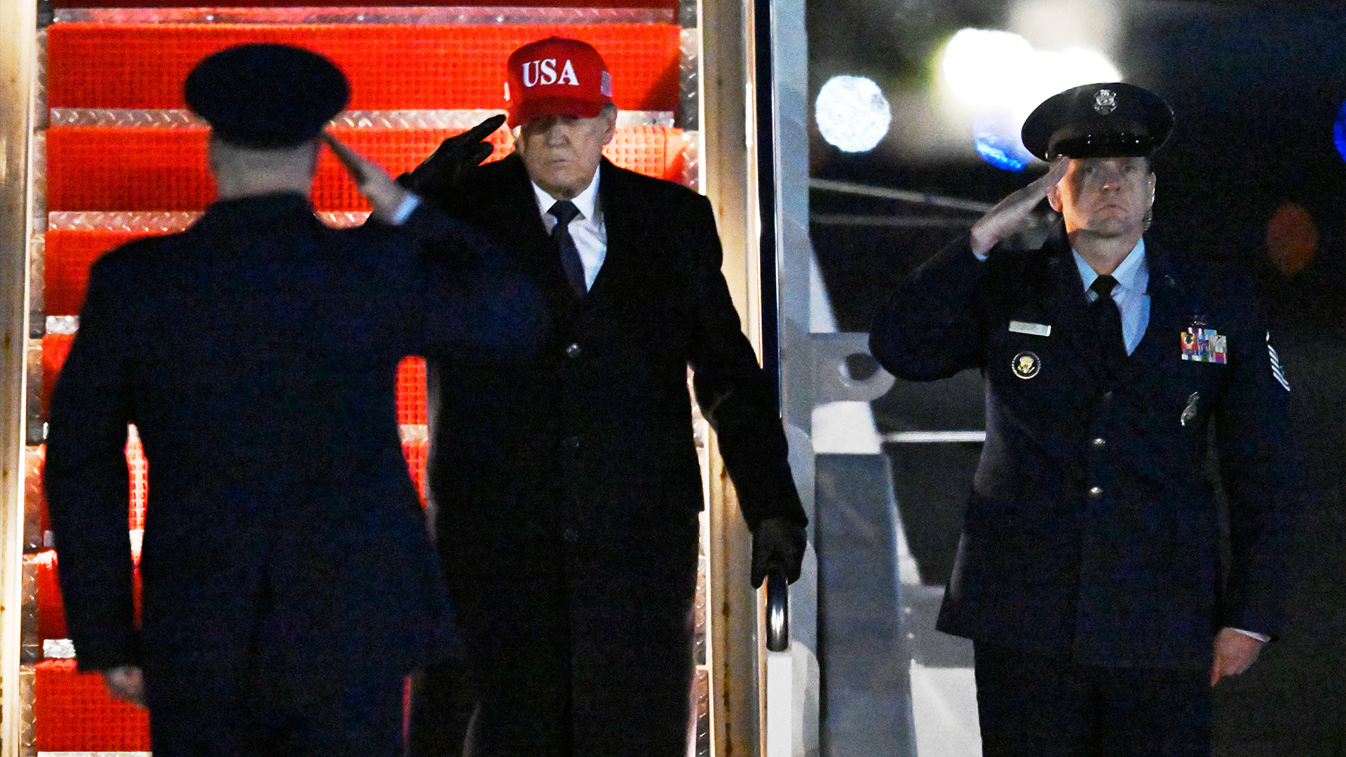 En man i mörk kostym stiger på eller av ett amerikanskt militärflygplan med uniformerad militär personal närvarande.