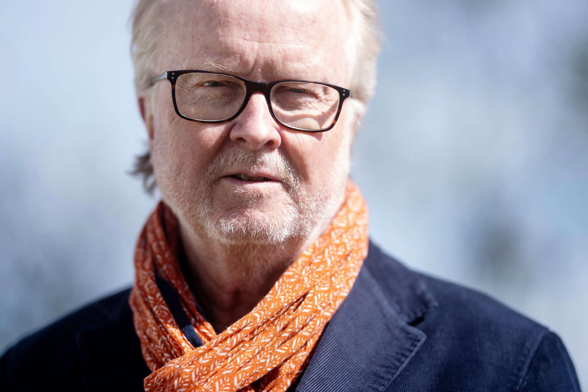 Porträtt av en äldre man med glasögon och orange halsduk mot en suddig bakgrund.