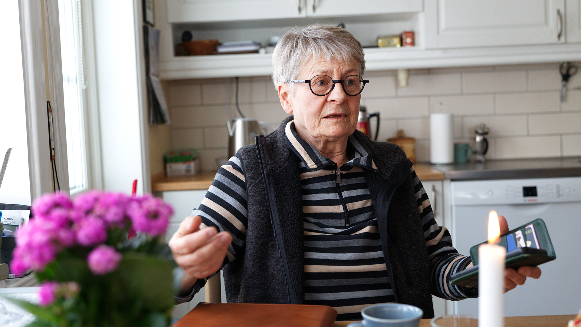 Äldre kvinna med glasögon sitter vid ett köksbord och gestikulerar medan hon talar. Rosa blommor och ett ljus står på bordet framför henne.