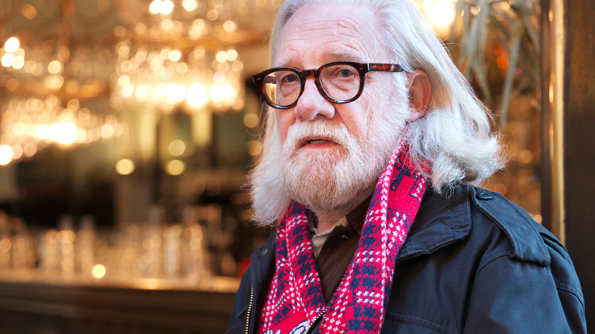 Porträtt av en äldre man med vitt hår, skägg och glasögon, klädd i mörk jacka och röd mönstrad halsduk.