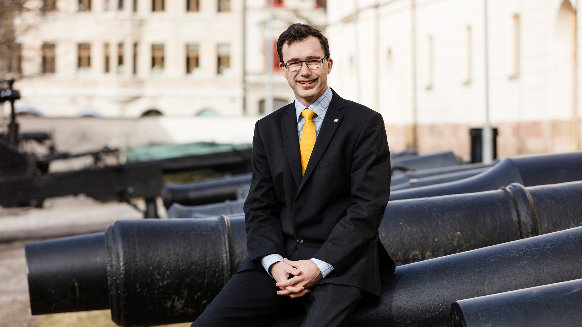 En man i mörk kostym och gul slips sitter på stora svarta rör utomhus. Han ler och tittar mot kameran. Byggnader syns i bakgrunden.
