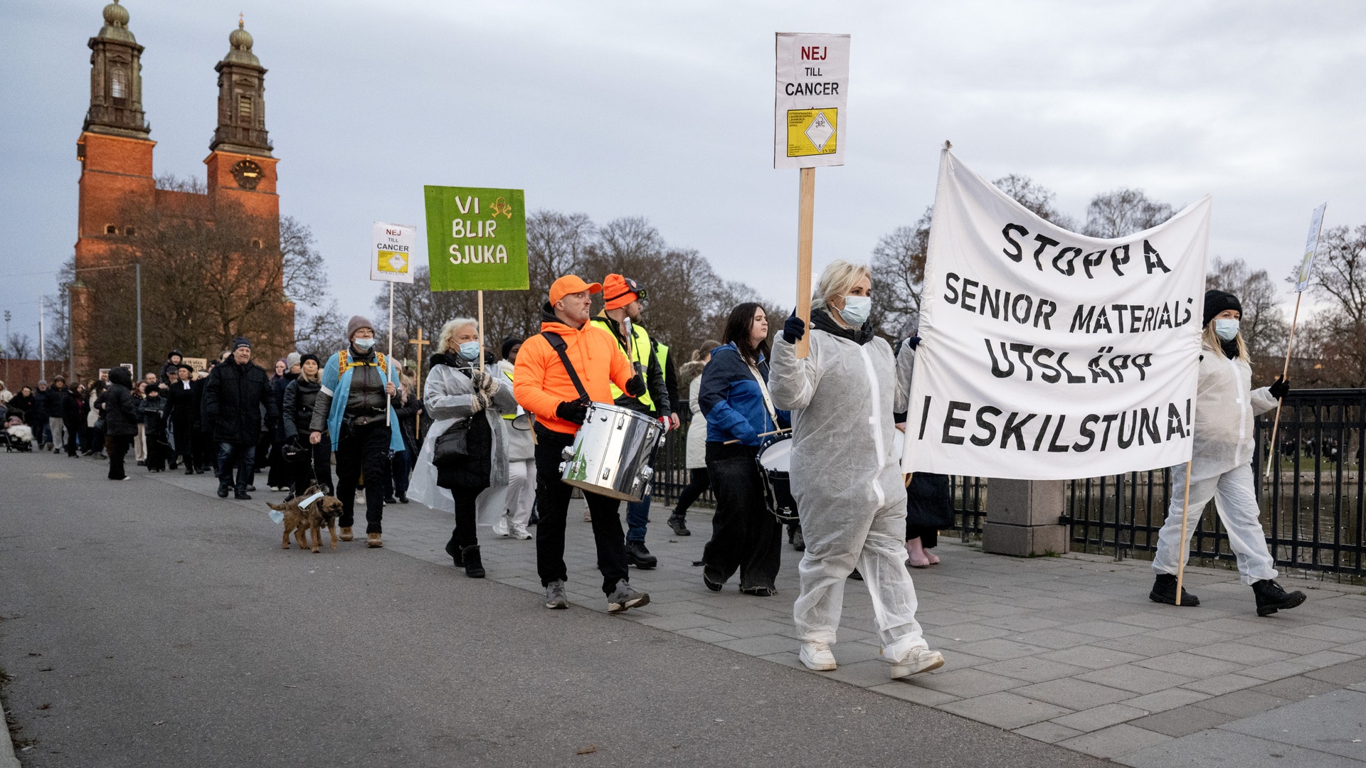 En grupp äldre människor demonstrerar på en öppen plats framför en röd tegelkyrka. De bär plakat med budskap på svenska och några är klädda i orange västar. Demonstrationen verkar handla om äldres rättigheter och välfärd.