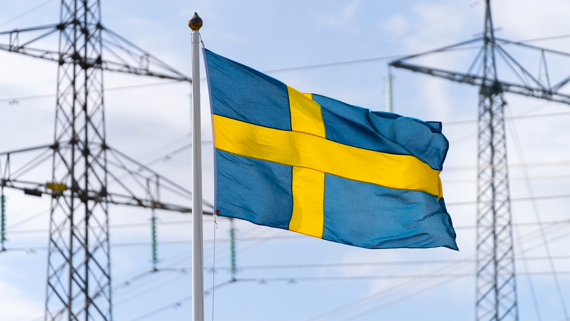 Svenska flaggan vajar på en flaggstång med kraftledningsmaster i bakgrunden.