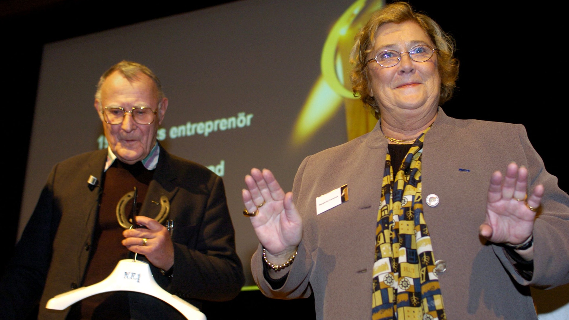 Två personer presenterar på en scen framför en skärm under ett evenemang.