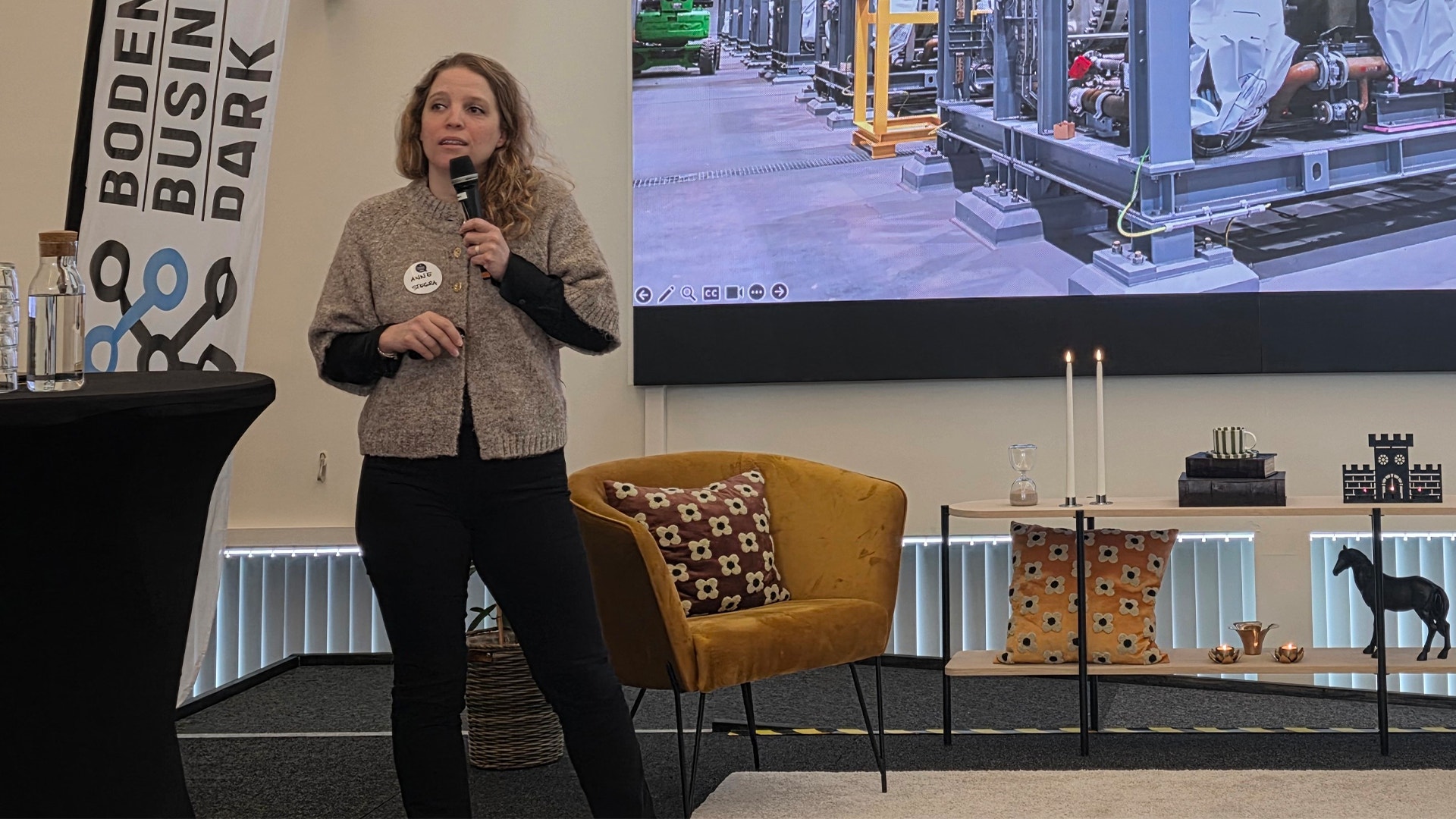 En kvinna presenterar vid ett evenemang, hon håller i en mikrofon framför en skärm som visar en stadsmiljö.