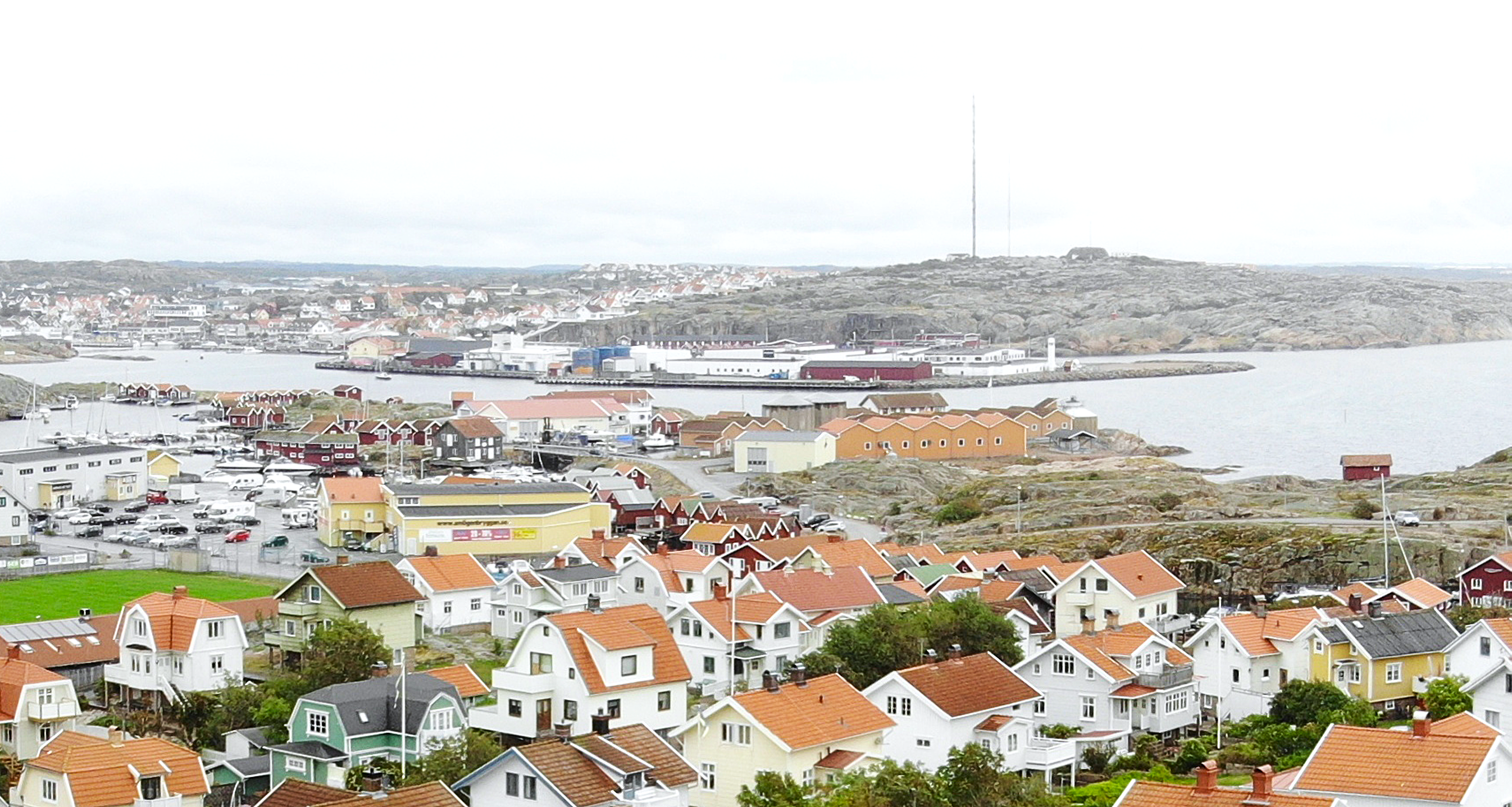 Luftfoto över Smögen med färgglada bostadshus med röda tegeltaken i förgrunden, hamn med båtar och industribyggnader längre bort, samt havet och kustlandskapet i bakgrunden.