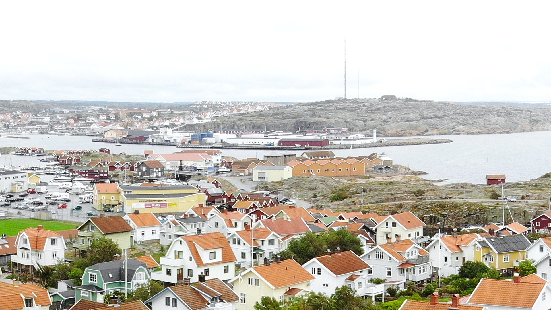 Luftfoto över Smögen med färgglada bostadshus med röda tegeltaken i förgrunden, hamn med båtar och industribyggnader längre bort, samt havet och kustlandskapet i bakgrunden.