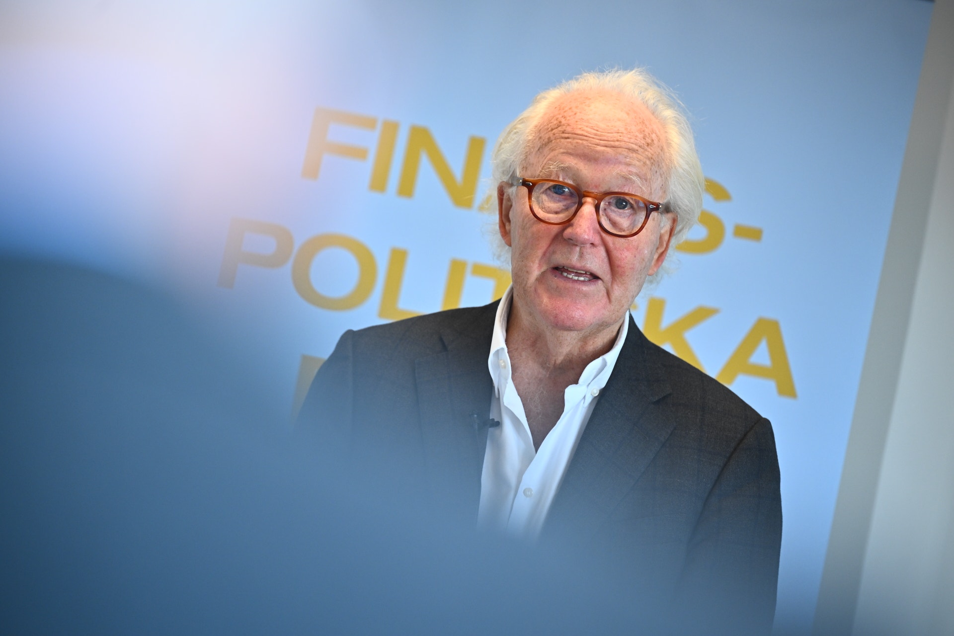 Äldre man med vitt hår och glasögon i mörkblå kavaj framför blå bakgrund med text om Finland och Polen.