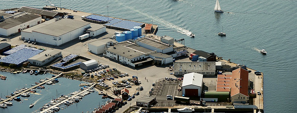 Flygfoto över en hamn med båtar, industriella byggnader och kajplatser vid vattnet.