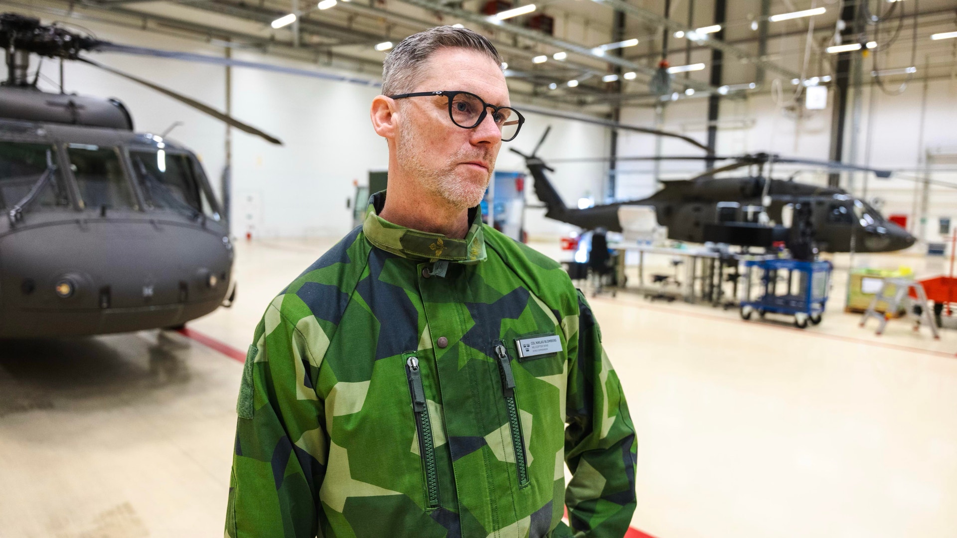Man i grön militär kamouflageuniform står i en helikopterhangar med militära helikoptrar i bakgrunden.