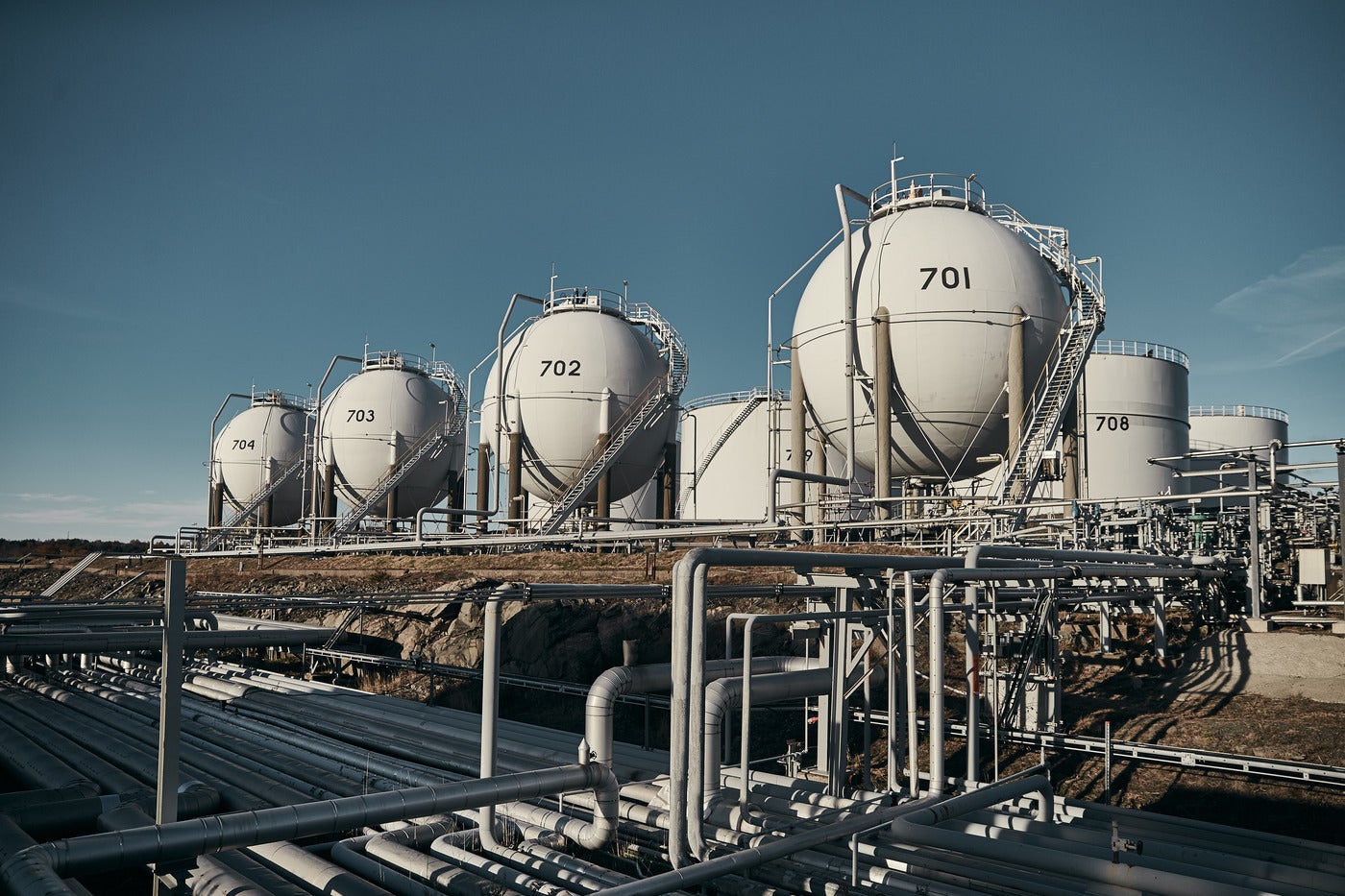 Industriell raffinaderi med flera vita lagertankar och rörsystem under klar blå himmel.